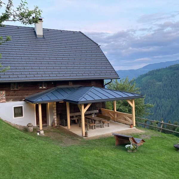 The farmhouse exterior includes a covered patio with wooden seating, an outdoor barbecue grill, and views of the surrounding mountains and forests.