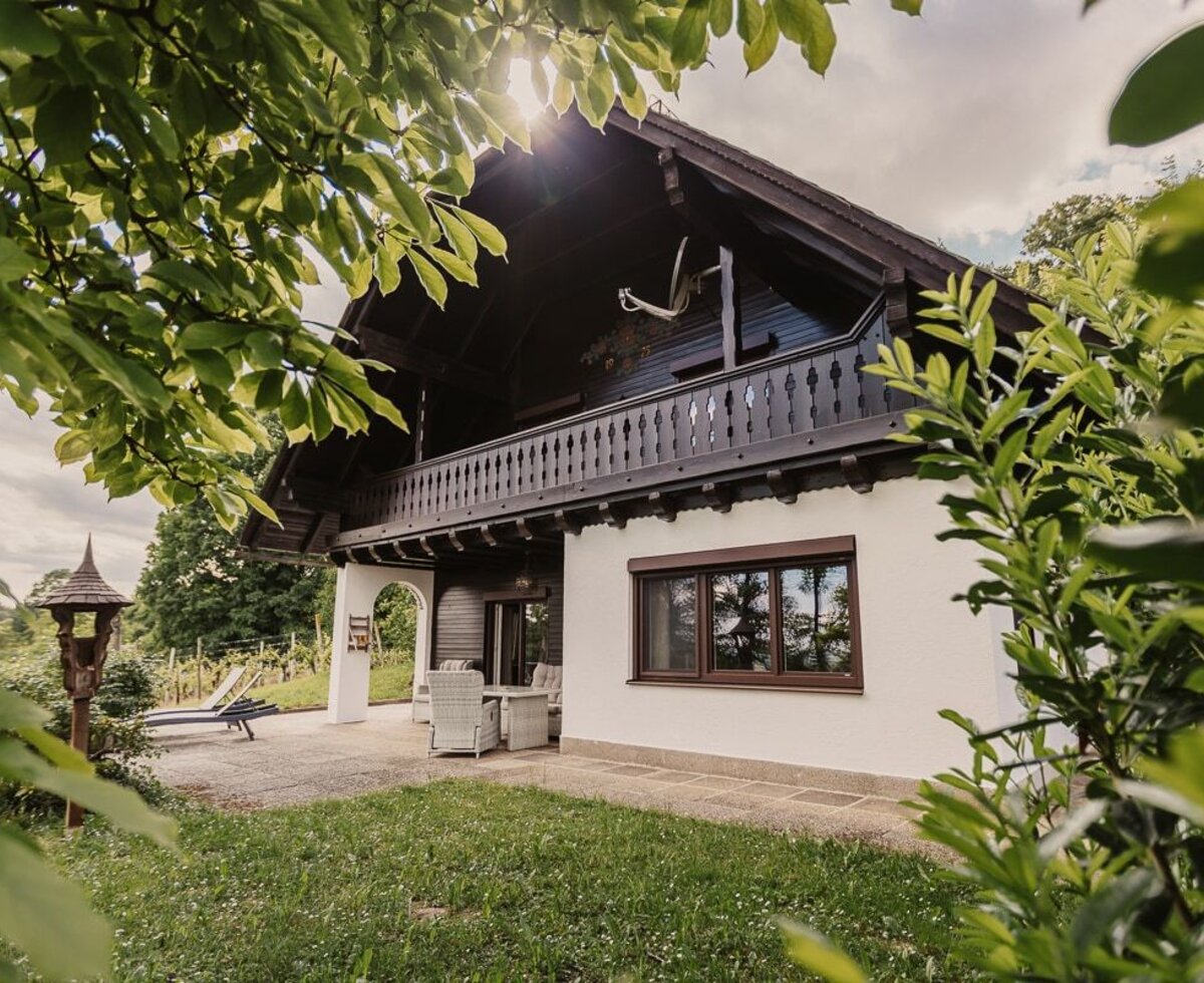 Der Bauernhof mit seiner weißen Fassade, dem Holzbalkon im Obergeschoss und der Terrasse mit Sitzgelegenheiten und Liegestühlen im Gartenbereich.