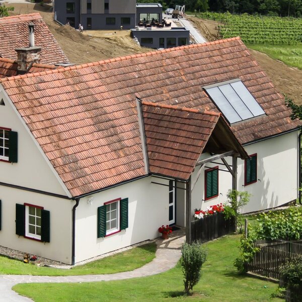 Außenansicht des Hofes mit weißer Fassade, roten Dachziegeln, grünen Fensterläden und einem Dachfenster, umgeben von einem gepflegten Garten mit Eingangsweg.
