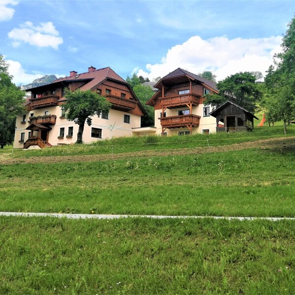 The farmhouse property features two main buildings with multiple balconies, a spacious lawn with a children's play area, and distant views of the Alps.