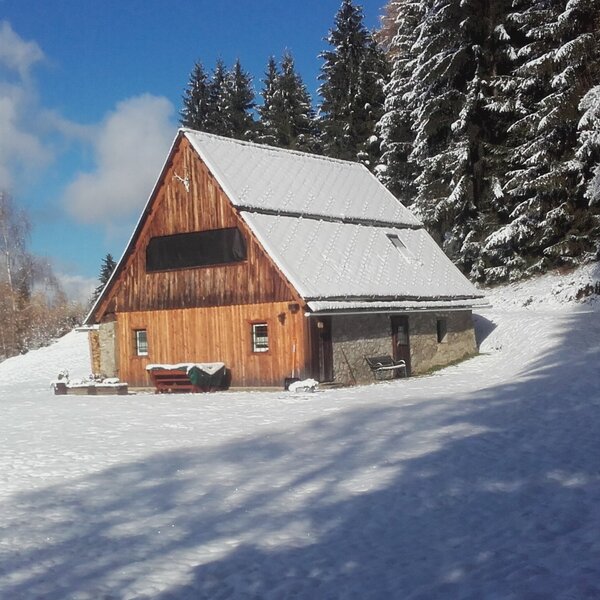 The farmhouse exterior in a snowy winter setting, featuring wooden and stone construction and surrounded by snow-dusted pine trees.
