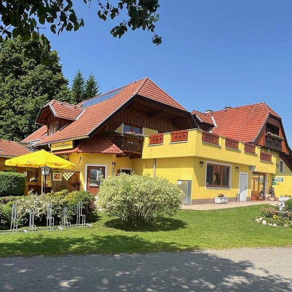 Exterior view of the farmhouse with a yellow facade, red roof, lawn, outdoor terrace with a yellow umbrella, and bike racks.