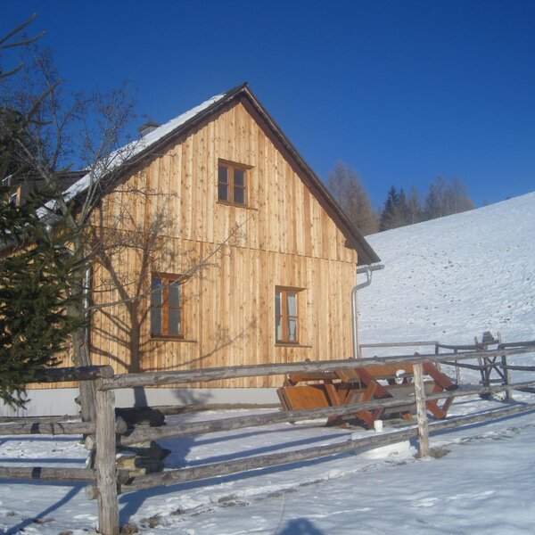 The wooden farmhouse exterior features a snow-covered roof, a wooden fence, an outdoor seating area, and a snowy hillside.