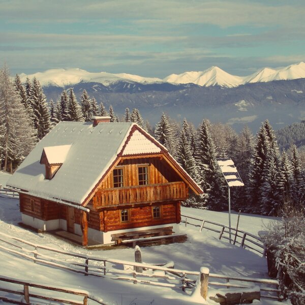 The wooden farmhouse exterior covered in snow, set amidst pine forests with distant snow-capped mountains.