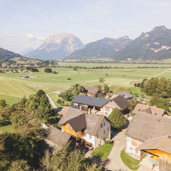 Ansicht des Bauernhofs in einem ländlichen Tal mit Feldern und den Bergen Grimming und Ennstal im Hintergrund.