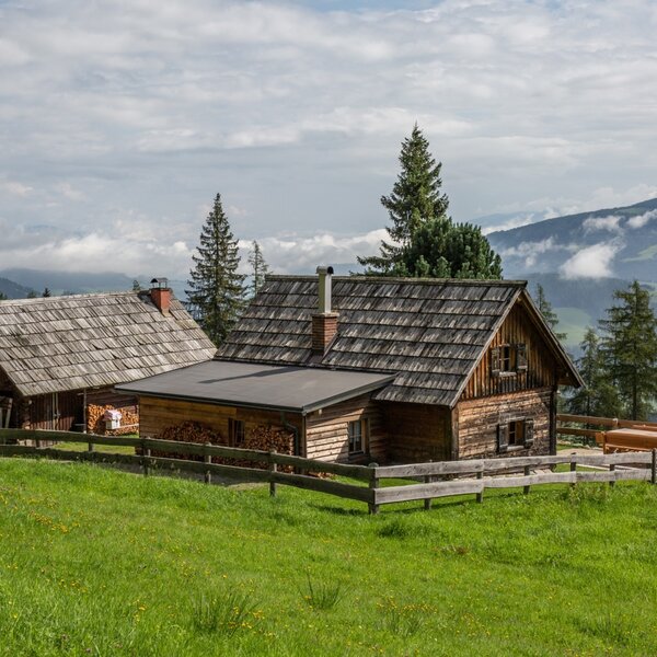 The wooden farmhouse and an adjacent building, featuring an outdoor seating area, are surrounded by a green lawn with mountain views.