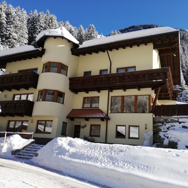 Der Bauernhof im Winter, mit Schnee auf dem Dach und den Balkonen, umgeben von schneebedeckten Wäldern.