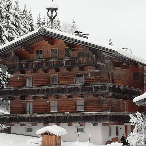 Der Bauernhof im Winter als mehrstöckiges Holzhaus mit Balkonen, umgeben von verschneiten Bäumen.