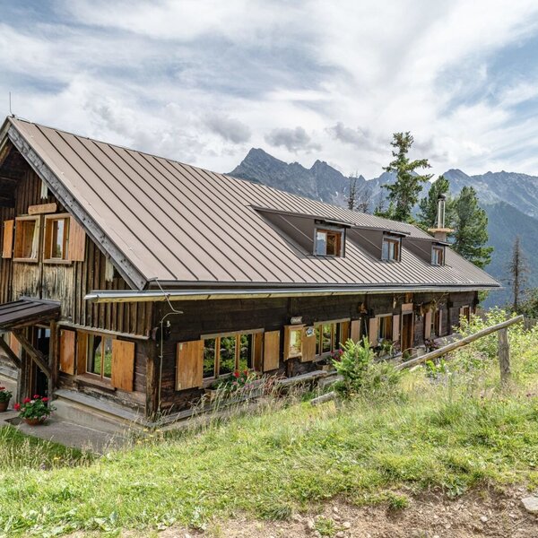 The Alpine Hut features a dark wooden facade with a metal roof and multiple windows with wooden shutters, set on a grassy slope with views of the surrounding mountains.