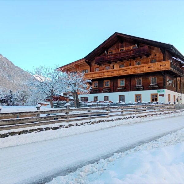 Der Bauernhof mit seiner traditionellen Holzfassade, umgeben von Schnee und der Berglandschaft.