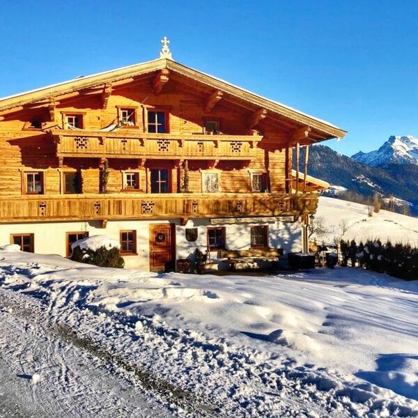 Außenansicht des Bauernhofs mit Holzfassade, Balkonen und schneebedeckter Umgebung vor Bergkulisse.