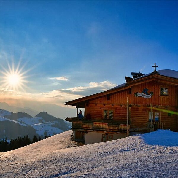 Die Alm, ein Holzgebäude auf einem verschneiten Berghang, mit Bergblick bei Sonnenuntergang.