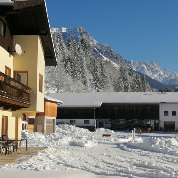 Der Bauernhof im Winter mit schneebedeckten Gebäuden, einer Terrasse und den Bergen im Hintergrund.