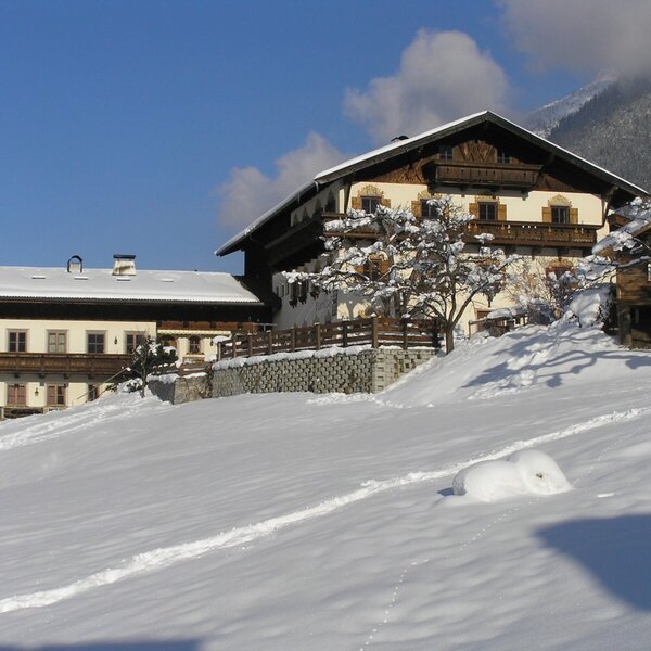 Der Bauernhof im Winter, mit seiner traditionellen Bauweise und der umliegenden, schneebedeckten Landschaft unter blauem Himmel.