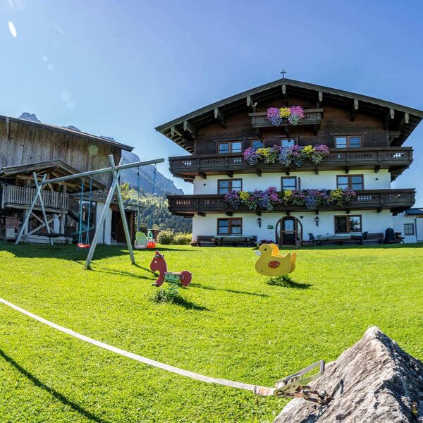 Vista esterna dell'agriturismo con balconi fioriti, un ampio prato con un'area giochi per bambini e una slackline.