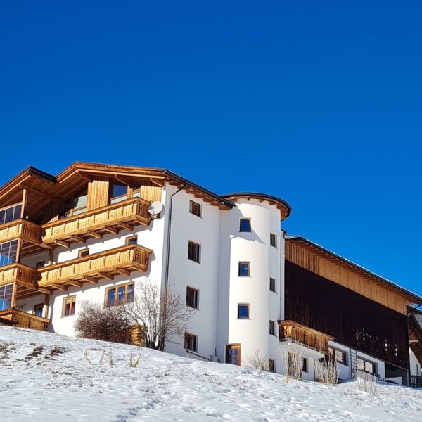 Der Bauernhof im Winter, mit weißer Fassade, Holzverkleidung und Balkonen, auf einem schneebedeckten Hügel.