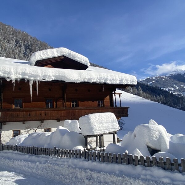 The farmhouse exterior with a snow-covered roof, icicles, and a balcony, set against a winter mountain landscape.