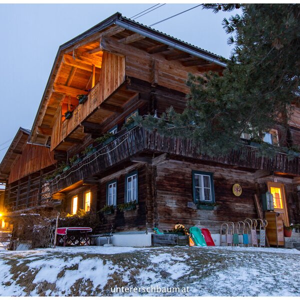 Der Bauernhof mit Holzfassade, Balkonen, beleuchteten Fenstern und Schlitten im verschneiten Außenbereich.