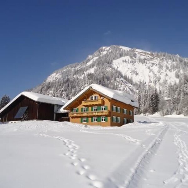 Der Bauernhof in einer verschneiten Winterlandschaft mit Bergen im Hintergrund.