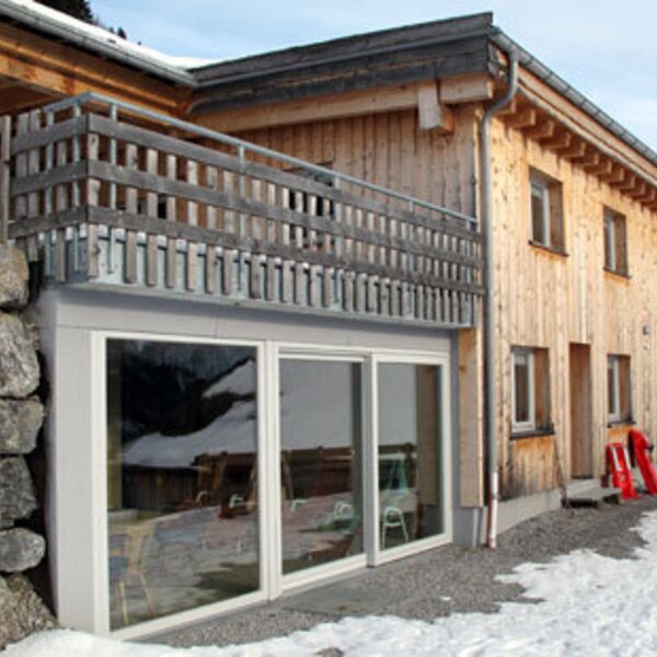 The wooden farmhouse exterior features a balcony and ground-floor glass doors, set against a snowy mountain environment.