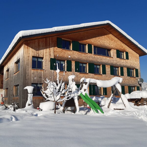 Außenansicht des Bauernhofs mit Holzfassade, grünen Fensterläden und schneebedecktem Dach, sowie einem teilweise sichtbaren Kinderspielplatz im Winter.
