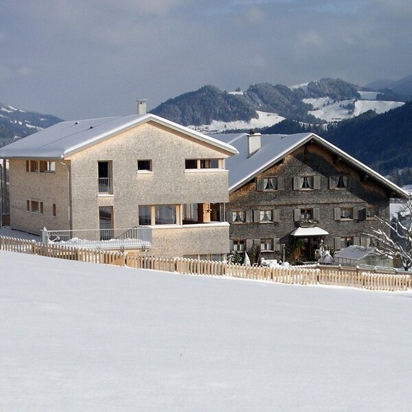Der Bauernhof präsentiert sich mit einem modernen Holzgebäude und einem traditionellen Bauernhaus, eingebettet in eine verschneite Berglandschaft.